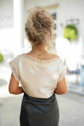Back view of the cream satin blouse showcasing the wide boat neckline framed by romantic, frayed eyelash lace that lays flat across the upper back. The model wears an updo hairstyle that highlights a dainty gold delicate chain necklace, with the satin fabric gently draped and tucked smoothly into a fitted black leather-look skirt.
