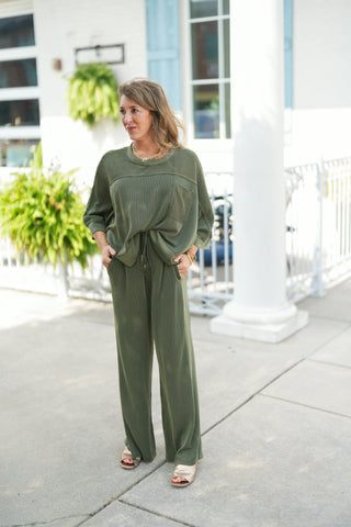 Full-length shot of a woman wearing a relaxed olive green two-piece ribbed knit lounge set. The top features a crew neck, subtle horizontal yoke seaming, a front patch pocket, 3/4-length drop-shoulder sleeves, and a front-hem tie accent. It is paired with matching wide-leg ribbed knit trousers featuring an elastic drawstring waistband and side slit pockets. Styled with metallic gold open-toe slide sandals, a gold link necklace, and gold bangles outdoors in front of a white building with light blue shutters.