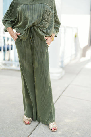 Close-up shot focused on the lower half of an olive green matching ribbed knit lounge set. Features high-waisted, wide-leg trousers crafted from a rich vertical-ribbed textured fabric, complete with side slit pockets and an adjustable drawstring waistband with gold aglets. Paired with open-toe metallic gold sandals outdoors.