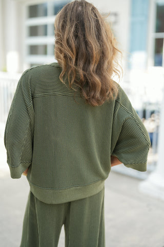 Back view of the olive green ribbed knit two-piece set showcasing the back yoke line, drop-shoulder elbow-length sleeves, and the fluid drape of the wide-leg pants. Highlights the full textured knit pattern and ribbed band hem for a cozy, elevated lougewear look.