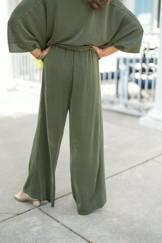 Back view focus on the olive green ribbed knit lounge pants, showcasing the relaxed high-rise fit, continuous vertical ribbed texture, and fluid wide-leg silhouette that flows down to the ankles. Features the model posing with hands on hips, highlighting the draping of the matching drop-shoulder top and gold block-heel sandals.