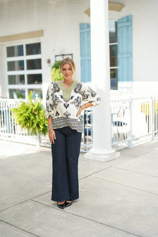 Full-length shot of a woman wearing a bohemian off-white tunic blouse with black and sage green damask-style floral printing. The blouse features a contrast sage green notched collar with rickrack trim, 3/4 sleeves with ruffled cuffs, and a patterned border hemline. Styled untucked over dark-wash wide-leg denim jeans and pointed-toe black patent leather heels outdoors in front of a white building with light blue shutters.