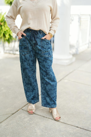 Woman modeling dark blue wide-leg denim jeans featuring an all-over tonal laser-etched floral pattern. The pants feature a comfortable high-rise waist with a dark blue drawstring tie, large utilitarian front patch pockets, and a relaxed wide-leg fit through the ankle. Styled with a light beige V-neck sweater and metallic gold open-toe low-block heel mules on a outdoor paved walkway.