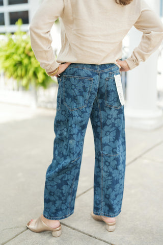 Back view showing the rear details of the dark blue floral print wide-leg jeans, showcasing two large back patch pockets, topstitching along the horizontal knee seams, and a relaxed straight-leg drape down to the ankles. The model stands with hands on her hips, wearing metallic gold mules and a beige pullover sweater tucked loosely into the waist.