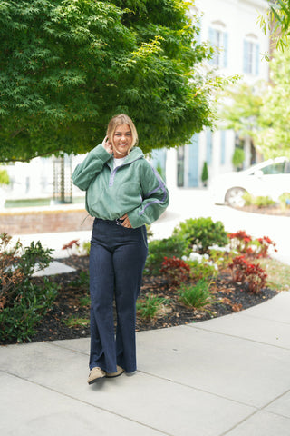 Woman posing outdoors under a leafy green tree wearing a sage green fleece half-zip pullover featuring lavender trim accents along the sleeves, collar, and front pocket contours. The oversized fleece jacket includes a adjustable toggle drawstring hem and front kangaroo pouch pocket, styled casually with dark blue wide-leg denim jeans and tan suede shoes on a paved sidewalk.
