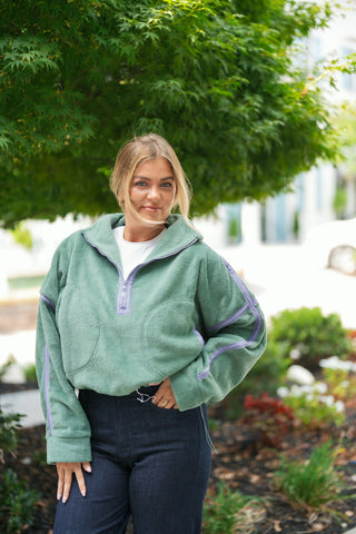 A portrait view highlighting the cozy plush texture of the sage green fleece half-zip pullover. Details the lavender piping along the half-zipper placket, stand collar, and drop-shoulder sleeve seams, layered over a white crew-neck tee and tucked slightly at the waist into dark blue jeans.