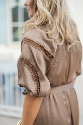Close-up side profile view highlighting the fine, subtle striped texture of the silky taupe fabric and the intricate tonal crochet lace embroidery along the raglan shoulder seams and flutter sleeves. Shows the gentle ruffle trim edging on the sleeve cuff and the fabric-matched tie belt at the waist.