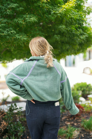 Back view of the sage green fleece pullover displaying the sporty raglan and drop-shoulder seam construction outlined with lavender trim. The model wears her hair in a messy low ponytail, showcasing the high fleece collar with a small back brand tag, paired with high-waisted dark blue jeans featuring clean back pockets.