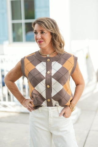 Medium close-up portrait highlighting the tailored construction and front details of the argyle button-up sweater vest. The textured knit top showcases decorative stitched argyle diamond patterns, a ribbed button placket with gold coin-style buttons, and a ribbed crew neckline, paired with a chunky gold chain necklace, gold bangles, and white jeans.