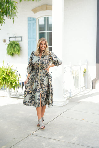 Full-length walking shot showcasing the fluid drape and hemline slit of the abstract printed midi dress in sage green, beige, and black. The smocked waistline defines the silhouette, while the exposed zipper front and puffed 3/4 sleeves add contemporary structure. Styled with grey peep-toe wedge sandals and gold jewelry on a bright paved walkway.
