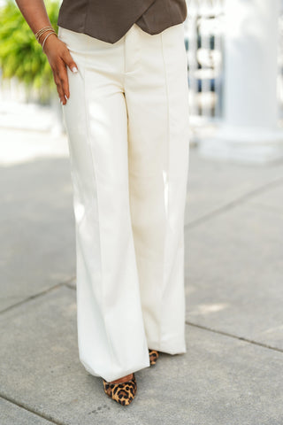 Lower-body shot of a woman modeling off-white high-waisted wide-leg trousers featuring sharp front pin-tuck leg seams, a zip fly front, and a floor-sweeping silhouette. The structured pants are paired with a dark olive-brown vest top featuring an asymmetrical overlap hem, styled with pointed-toe leopard print heels and delicate gold beaded bracelets on an outdoor paved walkway.