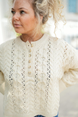 A close-up portrait highlighting the rich texture of the cream cable-knit henley sweater, detailing intricate openwork pointelle knit columns and braided cable patterns across the bodice. Focuses on the partial front placket trimmed with tortoiseshell-style buttons, a ribbed neckline, and a comfortable, relaxed fit.