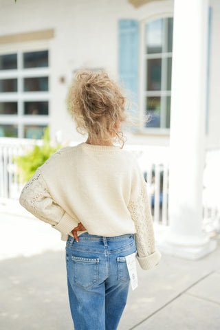 Back view showing the rear silhouette of the cream cable-knit sweater, featuring a smooth stockinette knit body contrasted by cable-knit textures along the sleeves. The model stands with hands on her hips, showcasing the slight cropped length that sits comfortably above the waist of high-rise medium-wash jeans with detailed stitching on the back pockets.