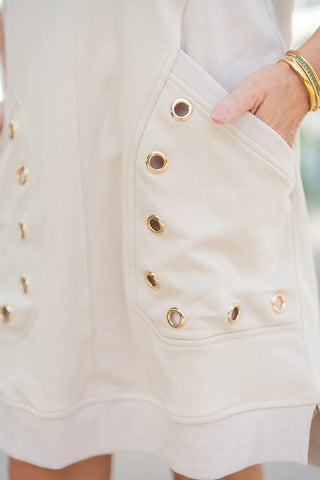 A detailed close-up shot highlighting the front patch pocket and side hem of the light beige sweatshirt dress. Focuses on the ribbed pocket lining and shiny gold metallic grommets arranged along the pocket border, showcasing the hand resting inside the pocket alongside a gold and emerald stacked bracelet.
