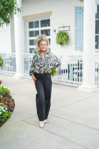 Full-length shot of a woman posing in a long-sleeve, relaxed-fit blouse featuring a swirling baroque-style abstract print in muted sage green, beige, black, and navy tones. The blouse is designed with a ruffled standing collar, a split V-neckline, and voluminous sleeves tucked into dark charcoal grey-black barrel leg denim jeans. Styled with off-white pointed-toe ankle boots, gold jewelry, and an updo hairstyle outdoors in front of a white building exterior with light blue shutters.