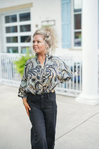 Woman posing outdoors wearing a silky sage green blouse featuring a bold black and beige scroll paisley print. The top features a ruffle stand collar, a open split V-neckline, flap chest pockets, and relaxed three-quarter sleeves. Tuck-styled into high-waisted washed black denim jeans and accessorized with gold hoop earrings and a delicate chain necklace in front of a white building background.