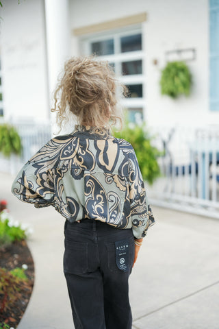 Back view of the sage green paisley printed blouse showcasing the straight back yoke line, ruffled stand collar detail, and relaxed drop-shoulder silhouette. Styled tucked into dark charcoal black jeans, viewed from behind with curly hair styled in a high messy bun.