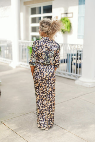 Back view of the abstract print shirt dress highlighting the continuous brushstroke pattern, back yoke seam detail, and straight maxi-length hemline. The model stands with her hair in a curly high updo, showcasing the tailored yet relaxed silhouette of the sleeves and skirt as she steps forward on a paved sidewalk.
