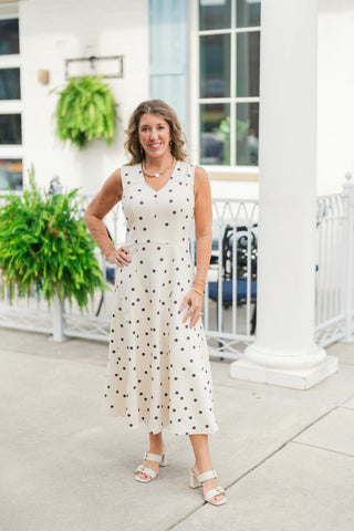 Woman posing in a sleeveless cream midi dress with black polka dots, featuring a soft V-neckline, structured shoulder inserts, and a fitted waist transitioning into a flowy A-line skirt. Styled for an elevated modern look with off-white double-strap block heel sandals, gold chain necklace, and gold bangles in front of a white architectural storefront patio.