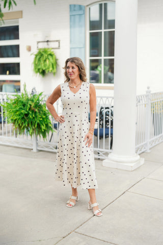 Full-length view of a woman wearing a cream and black polka dot A-line midi dress, showcasing its flattering cinched waistline, comfortable sleeveless design, and ankle-grazing hemline. The dress is paired with light cream block heel sandals with gold buckle accents for a timeless, sophisticated aesthetic.