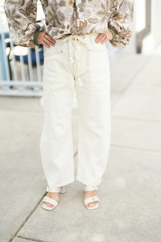 Woman modeling off-white wide-leg utility denim jeans featuring a braided rope drawstring waistband, front patch pockets, and contrast stitching along the seams. The high-waisted pants have a relaxed, straight-to-wide leg profile and are styled with an olive and beige floral blouse tucked into the waist, paired with off-white buckled block-heel sandals on a concrete walkway.