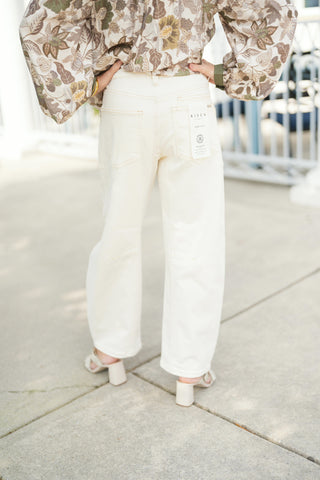 Back view detailing the rear fit of the off-white wide-leg utility jeans, showcasing two large back patch pockets, a fitted high-rise waist, and subtle horizontal knee seam details. The model stands with hands on her hips, showing the wide leg cut extending over off-white block-heel mules, paired with an olive green and tan floral long-sleeve blouse with elastic cuffs.