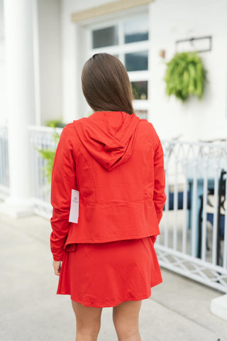 Back view showing the rear details of the bright red activewear set, featuring a structured hood, subtle raglan shoulder stitching, and an elasticized waist hem on the jacket. The model stands facing away, highlighting the comfortable drape of the lightweight jacket and the fluid A-line coverage of the matching red athletic skort.