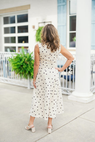 Back view of the sleeveless cream polka dot midi dress displaying a clean high-back neckline with a subtle center seam, structured waist paneling, and a sweeping A-line skirt. Shown paired with off-white chunky block heel sandals and styled in an outdoor setting.
