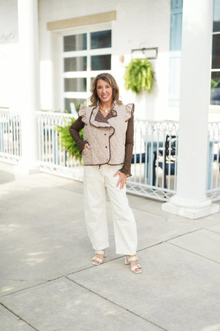 Full-length portrait displaying the reversible quilted vest worn on the light beige side featuring a brown ditsy floral print. Details highlight the brown piped trim, front patch pockets, ruffled shoulder accents, and the contrasting polka dot inner lining displayed at the open collar. Styled with the sheer brown long-sleeve base layer, cream casual trousers, and neutral block-heeled sandals.