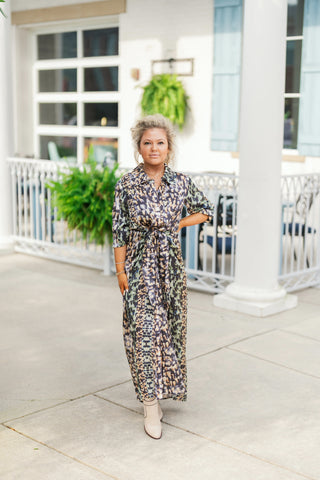 Woman posing outdoors in an abstract print ankle-length shirt dress featuring an all-over muted multi-color brushstroke pattern in shades of black, tan, sage green, and cream. The lightweight dress features a classic pointed collar, three-quarter sleeves with rolled cuffs, a button-down placket, and a fabric self-tie sash at the waist. Styled with cream pointed-toe ankle boots, gold hoop earrings, and a dainty gold necklace against a bright building backdrop.