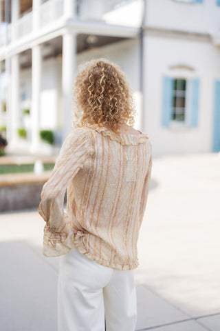 Back view of a blonde woman wearing the sheer yellow-and-peach vertical striped ruffle blouse, showcasing the subtle ruffle trim along the collar and hemline, as well as the fluid, relaxed drape over white trousers.