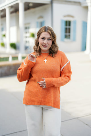 Three-quarter portrait view of a woman smiling in an orange crewneck sweater with contrast white stitching accents across the shoulders. Highlights the relaxed knit silhouette, long ribbed cuffs, and gold jewelry accents including rings and a cross pendant.
