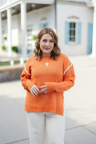 Three-quarter portrait view of a woman smiling in an orange crewneck sweater with contrast white stitching accents across the shoulders. Highlights the relaxed knit silhouette, long ribbed cuffs, and gold jewelry accents including rings and a cross pendant.