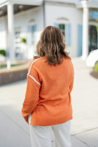 Back view of a woman in an orange marled knit pullover sweater and off-white wide-leg pants. Showcases the smooth back knit construction, white blanket stitching running down the back shoulder seams, and the stepped side slit hemline.
