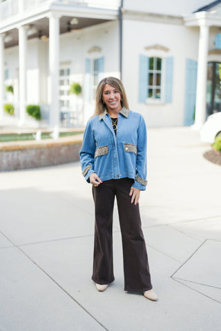 Woman standing outdoors wearing a light wash blue denim jacket featuring contrasting leopard print fabric on the pointed collar, front flap chest pockets, and turned-up cuffs. The jacket has raglan sleeves and a relaxed, straight hemline. Styled with dark brown wide-leg pants and pointed light beige ankle boots in front of a white building background.