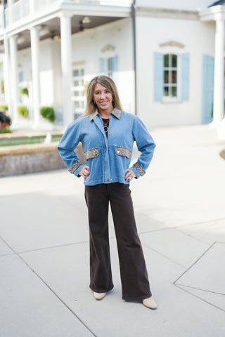Woman posing outdoors in a light wash blue denim jacket featuring animal print leopard accent trim on the collar, cuffs, and front chest patch pockets. Styled layered over a black ribbed top and paired with dark chocolate brown high-waisted wide-leg pants and pointed cream boots in front of an elegant white building backdrop.