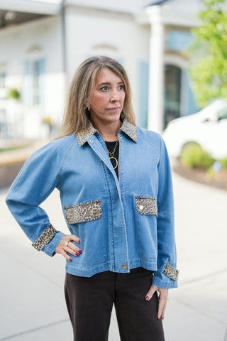 A detailed upper-body view highlighting the leopard print fabric accents on the collar, front utility pockets with gold snap button closures, and cuff trims of the light blue denim jacket. Styled layered over a black top with a chunky gold chain link necklace.