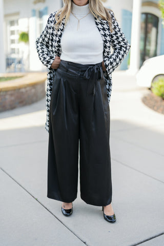 Close-up front view of the Black Satin Wide-Leg Trousers highlighting the silky fabric lustre, structured waistband with belt loops, self-tie bow accent, and deep front pleating. Shows the relaxed drape down to the wide ankle hem paired with black pumps.