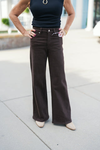 A detailed shot from the chest down highlighting the high-rise waist and smooth front construction of the dark brown wide-leg pants. Displays a zip fly with silver button closure, five-pocket denim details, and full-length wide trouser legs draped over cream pointed-toe boots beneath a fitted black sleeveless top.