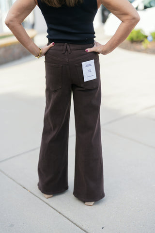 Back view from the waist down showcasing the fit and rear coverage of the chocolate brown Good American Palazzo Ankle wide-leg pants. Highlights classic back patch pockets with brand tag detailing, a high-rise waistband fit, and a wide-leg hemline draped over cream boots.
