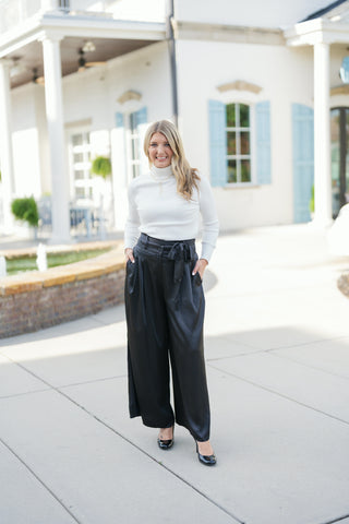 Woman posing outdoors in the Black Satin Wide-Leg Trousers, a high-waisted fluid pair featuring a matching tie-belt sash, tailored waist button, pleated front, and side slant pockets. Styled for an elegant, elevated look with a fitted white ribbed turtleneck sweater, black patent leather pointed heels, and minimal gold jewelry in front of a white building background.
