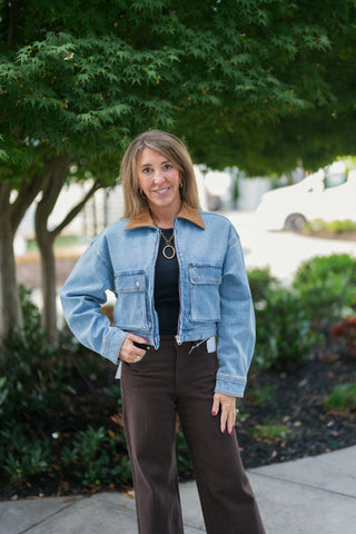 A mid-length front view highlighting the cropped hem and drop-shoulder relaxed fit of the light blue denim jacket with its warm brown collar contrast. Paired with dark brown wide-leg trousers, a black ribbed shirt, and chunky gold jewelry.