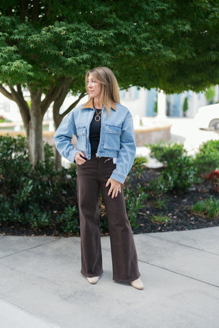 Woman posing outdoors in a light wash blue cropped denim jacket featuring a contrasting tan corduroy collar, zip-front closure, and dual chest flap pockets. Layered open over a fitted black ribbed top and paired with dark chocolate brown high-waisted wide-leg pants and cream boots. Styled with a gold ring pendant chain necklace in front of a green tree backdrop.