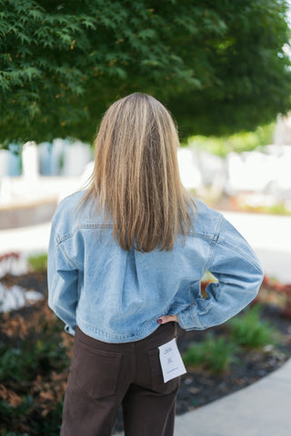 Back view showcasing the back yoke seam, drop shoulders, and cropped waistband of the light blue denim jacket. Highlights the fit and high-rise waistband of the chocolate brown Good American wide-leg pants with back patch pocket detail.