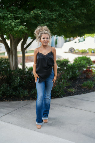 Woman posing outdoors in a black satin camisole top featuring delicate eyelash lace trim along the V-neckline and asymmetrical hemline. The top is paired with medium-wash bootcut jeans featuring bold cuffed hems, styled with tan square-toe boots, gold hoop earrings, and a high curly updo in front of a lush green tree.