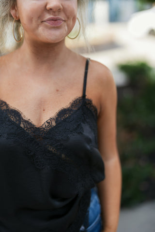Portrait shot focusing on the neckline of a black satin camisole, highlighting the detailed eyelash lace overlay along the delicate V-neck, thin shoulder straps, and subtle sheen of the fabric. Styled with gold hoop earrings.