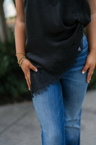 Close-up shot showing the asymmetrical sweeping hemline of a black satin top accented with continuous floral lace trim against medium-wash blue jeans. Showcases the intricate eyelash fringe edge and delicate hand jewelry.