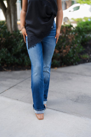 A detailed front shot from the waist down highlighting the medium-wash denim texture, subtle whiskering across the lap, and slim fit through the thighs. Shows the wide cuffed hems resting over tan leather square-toe boots, draped beneath the scalloped lace edge of the black satin top.