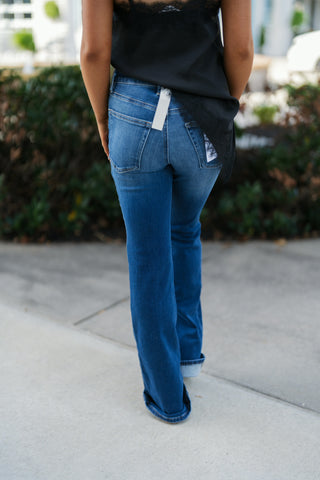 Back view from the waist down showcasing the fit and rear coverage of the medium-wash bootcut jeans. Details classic back patch pockets, a fitted waist, straight-to-bootcut leg line, and broad folded cuffs above tan boots, worn under the flowing black satin and lace top hem.