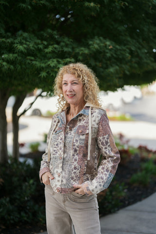 Woman posing outdoors in a button-front patchwork denim-style jacket featuring an intricate mix of floral, paisley, and earth-toned bohemian prints in beige, rust, and slate blue. The structured jacket includes a cream knit contrast drawstring hood, copper-tone metal buttons, chest flap pockets, and is styled with neutral beige pants.